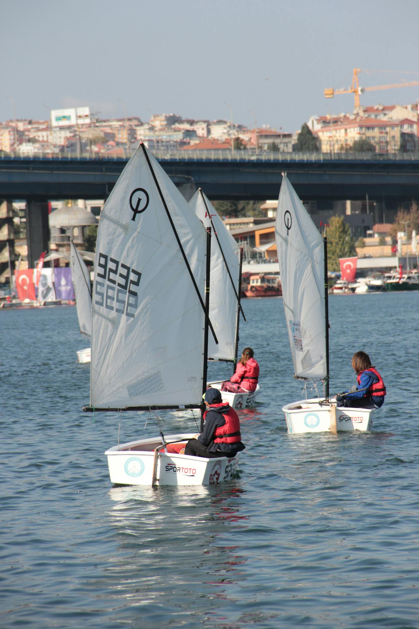 Children navigating sailboats in a calm urban waterway near a bridge.